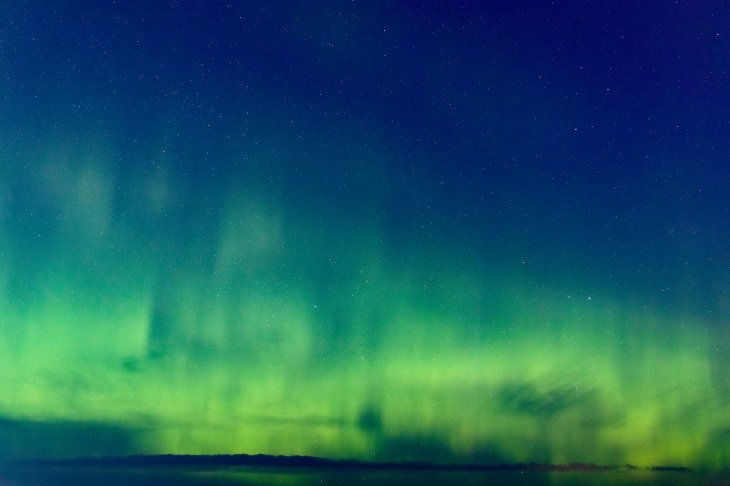 View of a clear night sky over a lake. Hundreds of stars are visible in the top half of the photo, emerald green aurora borealis cover the sky from left to right. 